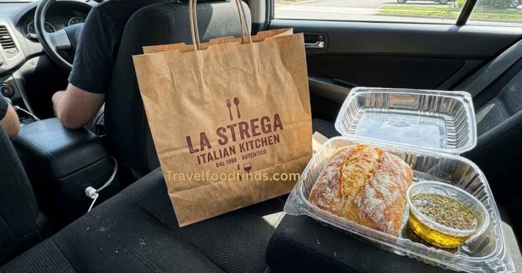 A takeout bag from an Italian restaurant with a container of complimentary crusty bread and herb olive oil dip.
